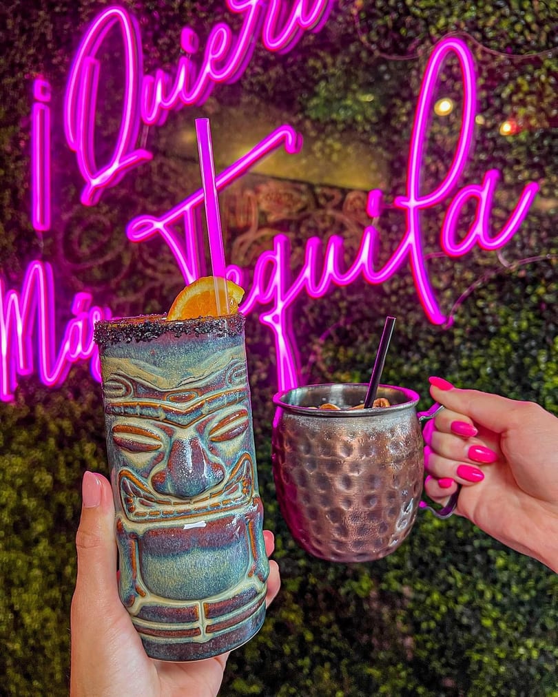 Two hands holding cocktails, a tiki mug and a copper mug, in front of a pink neon sign.