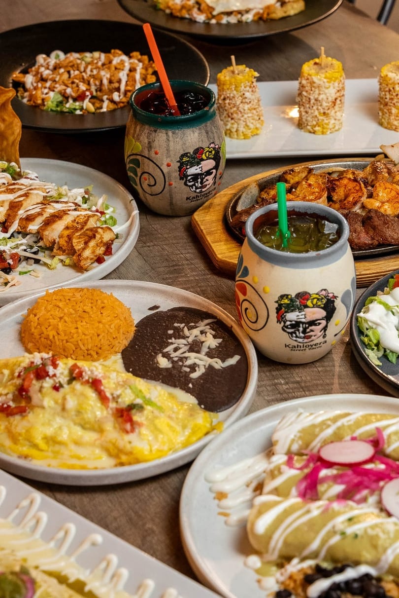 A table laden with various Mexican dishes, including fajitas, enchiladas, and drinks in Kahlolera mugs.