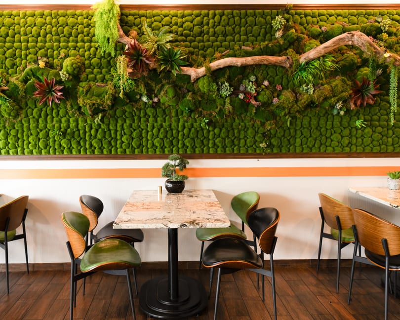 Restaurant interior with a green moss wall, marble-top tables, and modern chairs with green and black seats.