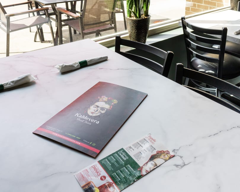 A Kahlovera Street Tacos menu and drink list on a white marble table with outdoor seating visible.