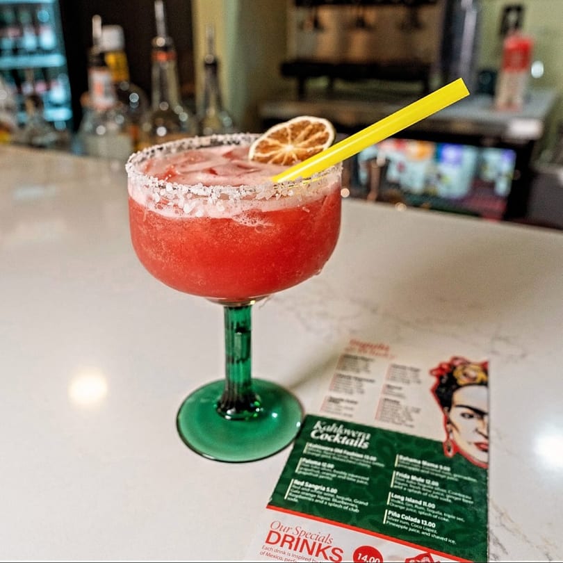 A vibrant red margarita with a salted rim, lime, and yellow straw sits on a bar next to a menu.