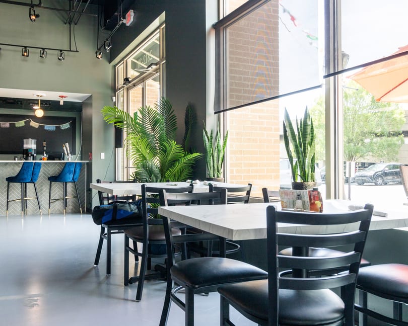 Modern restaurant interior with marble tables, black chairs, a bar with blue stools, and large windows.