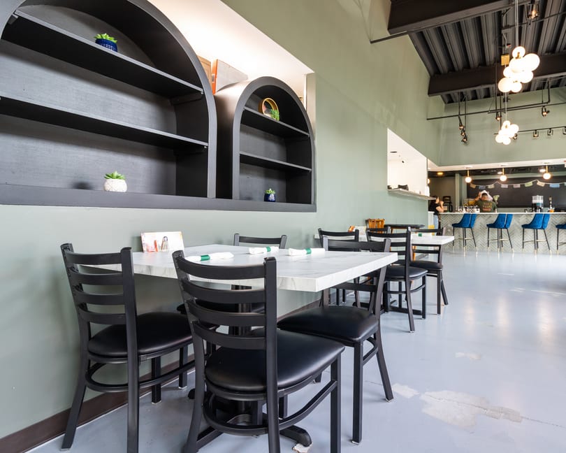 Restaurant interior with black chairs, white marble tables, and a bar with blue stools in the background.