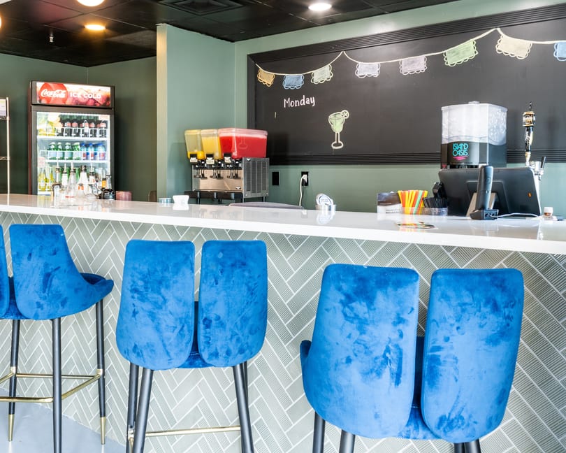A modern bar area with blue velvet stools, a white counter, and a chalkboard menu.