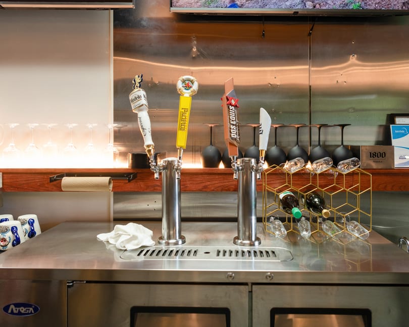 A bar counter with beer taps, wine bottles in a gold rack, and various glasses.
