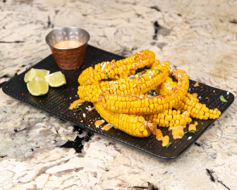Elote-style corn on the cob with sauce, cheese, and lime wedges on a black platter.