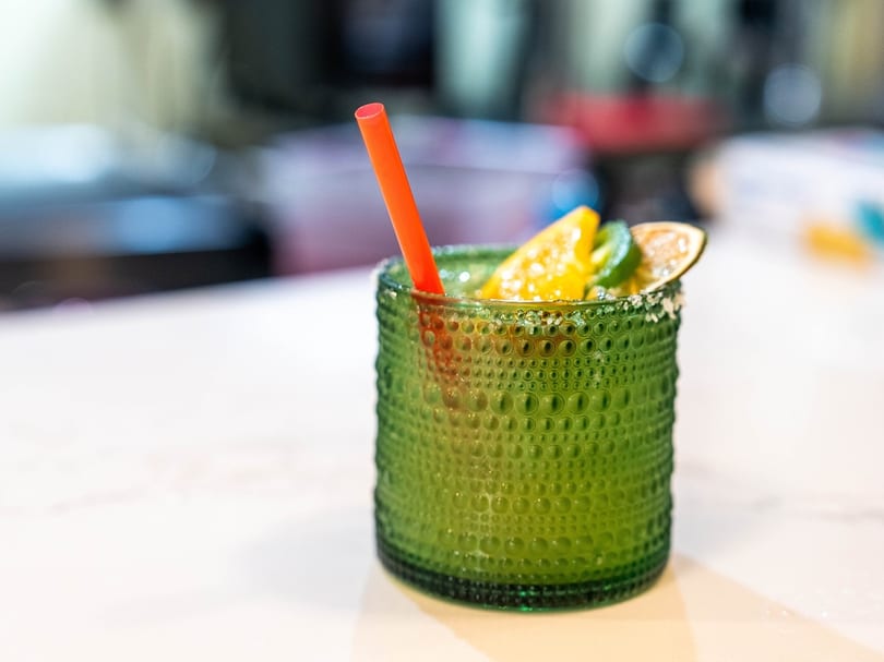 Green textured glass with a margarita, garnished with citrus slices, a jalapeño, and a red straw.