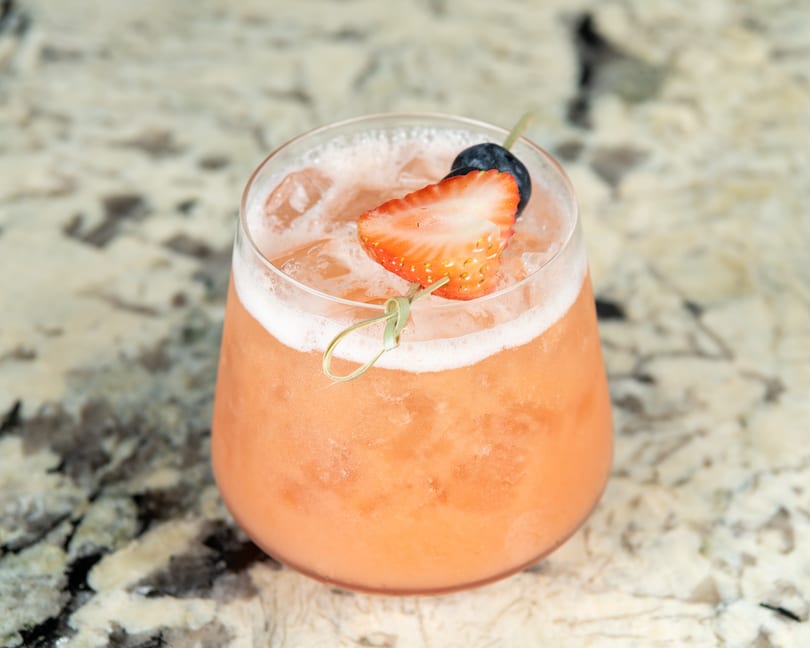 A refreshing pink cocktail with ice, garnished with a strawberry half and a blueberry on a skewer.