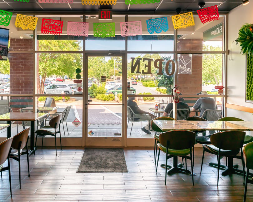 Restaurant interior with colorful papel picado banners, tables, chairs, and patrons visible through large windows.