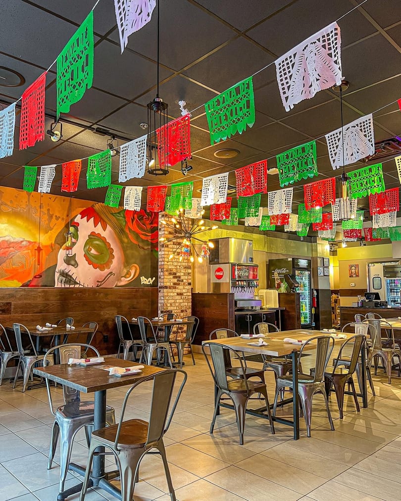Vibrant Mexican restaurant interior with colorful papel picado, a large mural, and industrial-style tables and chairs.