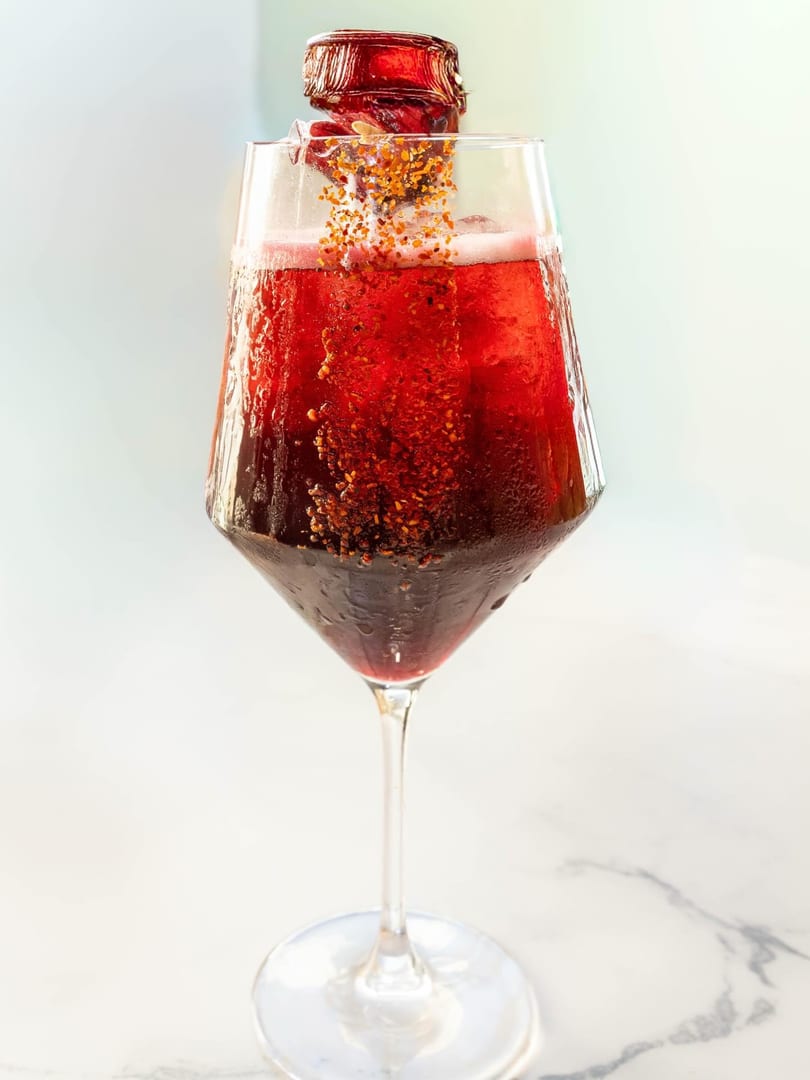 Red cocktail in a wine glass with a bottle pouring in golden sprinkles.