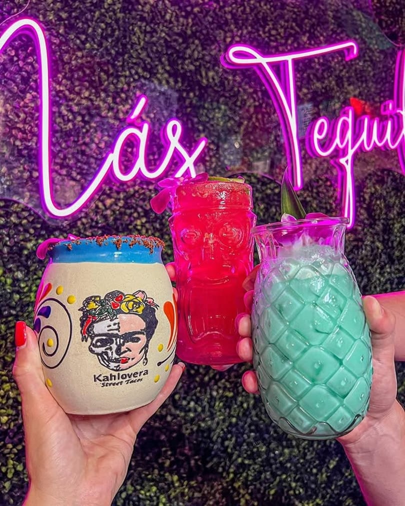 Three colorful cocktails, including a Frida Kahlo mug, held against a neon-lit green wall.