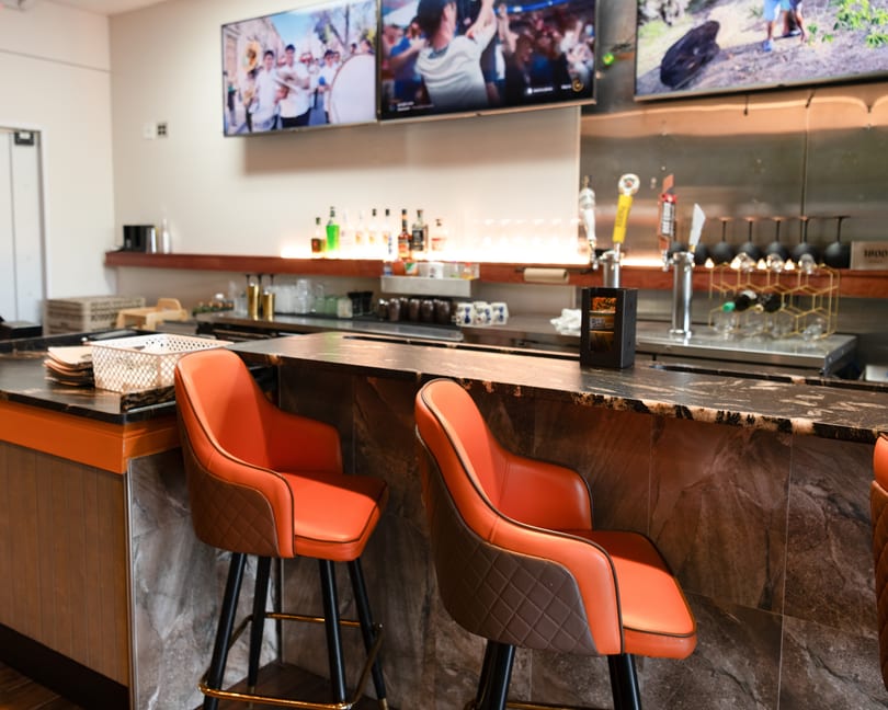 Empty bar area with orange bar stools, a dark marble counter, and TVs displaying sports.