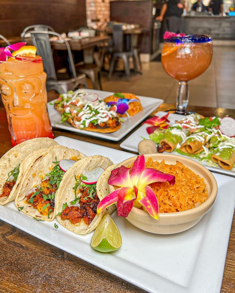 Tacos, rice, and other Mexican dishes with cocktails on a wooden table in a restaurant.