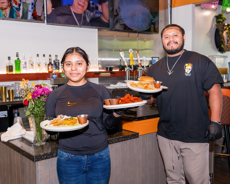 Two restaurant staff members smiling, holding plates of food including tacos, wings, and a burger with fries.