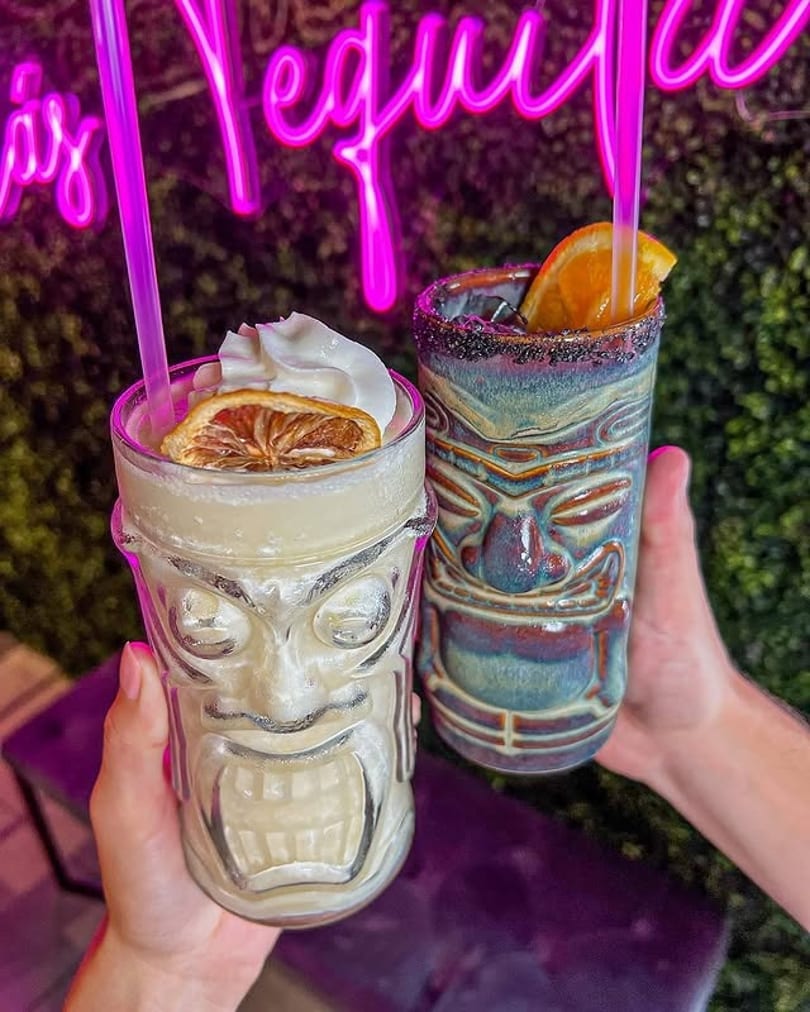 Two hands hold tiki drinks with garnishes, against a neon pink sign and green foliage.