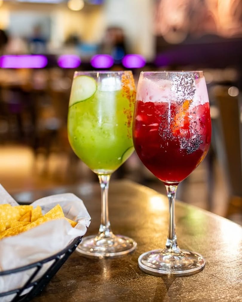 Two colorful cocktails, one green and one red, with a basket of tortilla chips on a table.