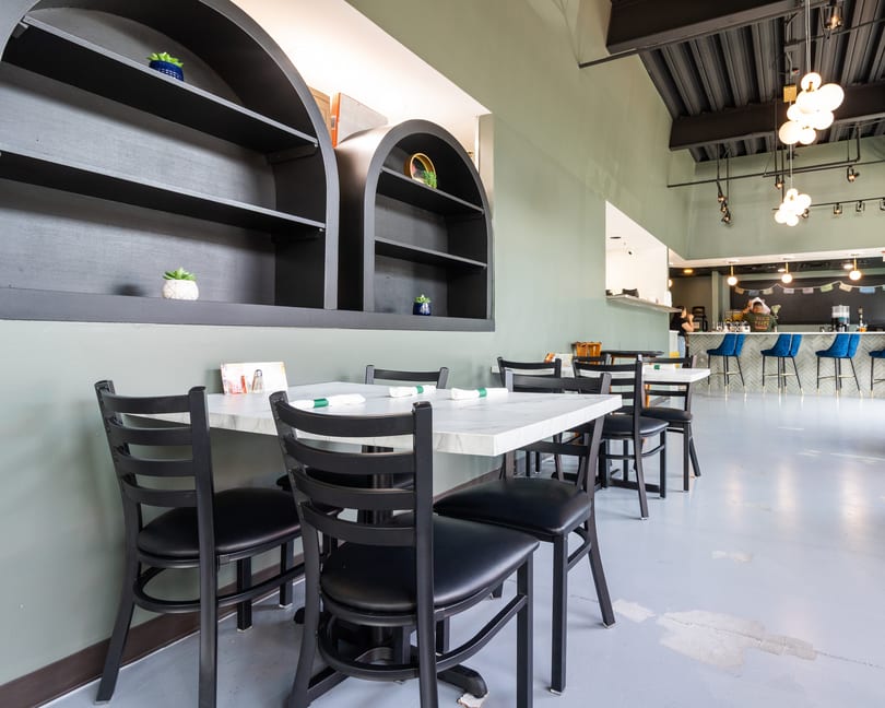 Restaurant interior with black chairs, white marble tables, and a bar with blue stools in the background.