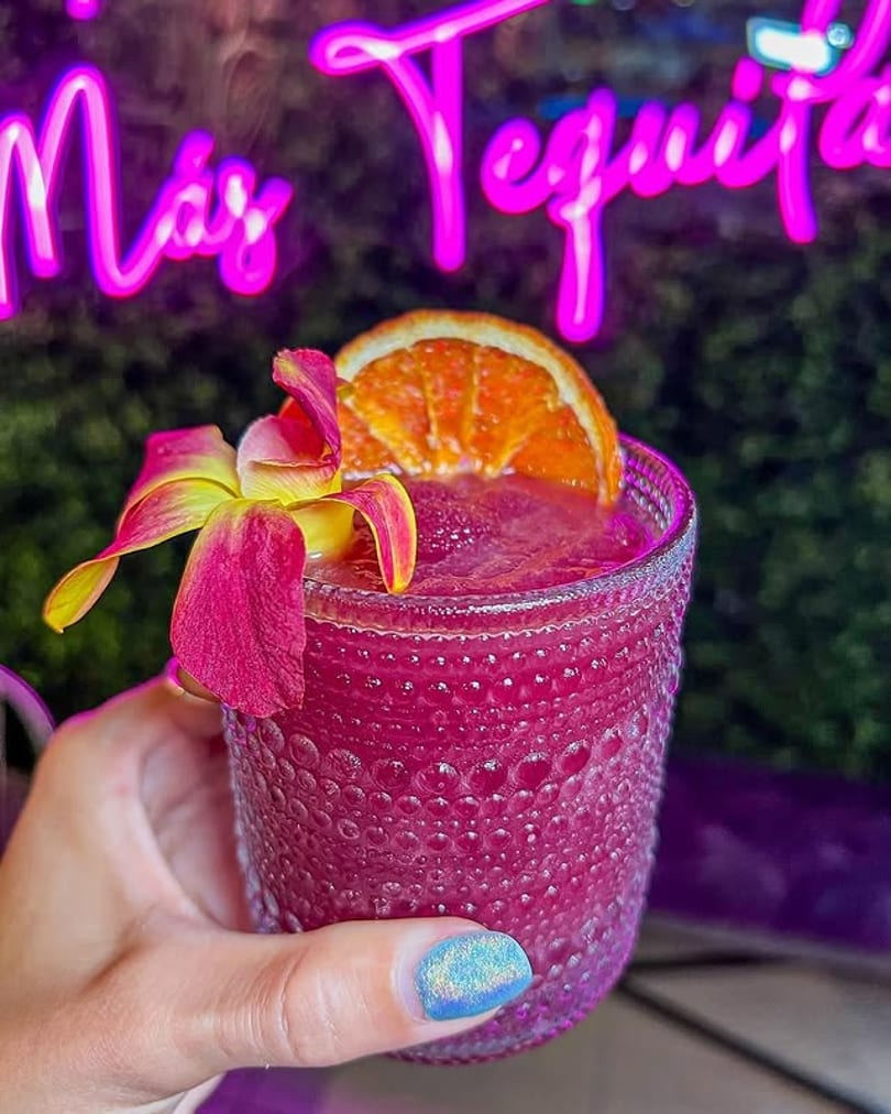 Hand holding a vibrant pink frozen drink garnished with an orange slice and orchid, under a neon sign.
