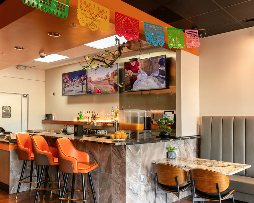 A vibrant restaurant interior with a bar, orange bar stools, and colorful papel picado decorations.