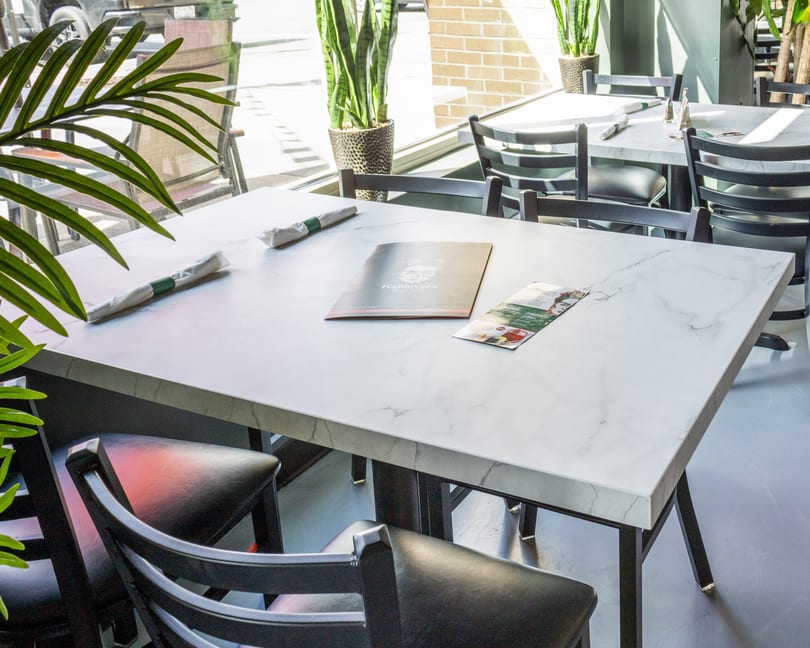 A clean, empty restaurant table with a white marble top, black chairs, and menus, bathed in sunlight.