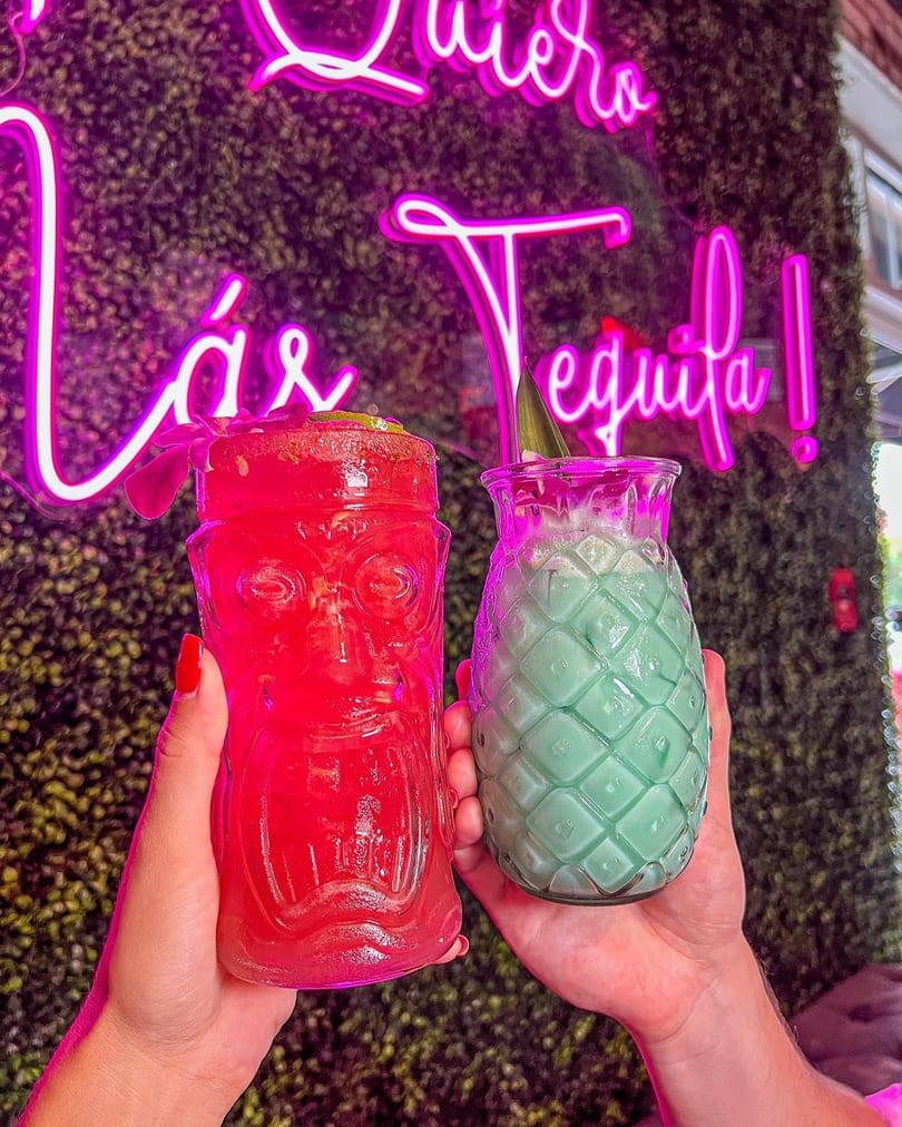 Two hands hold a red tiki drink and a green pineapple drink against a neon sign.