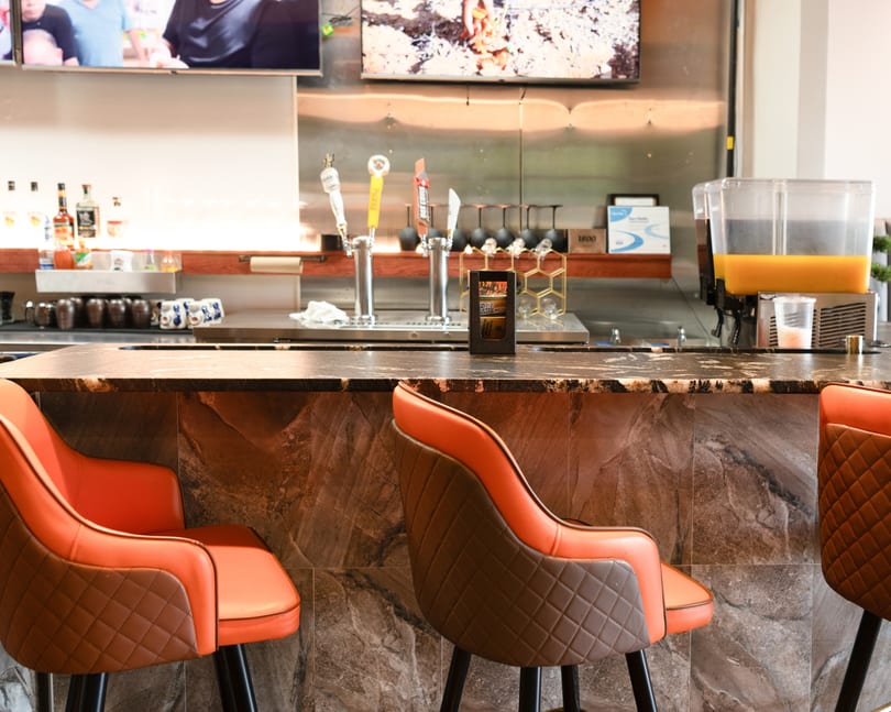 A modern bar with orange and brown bar stools, beer taps, and a juice dispenser.