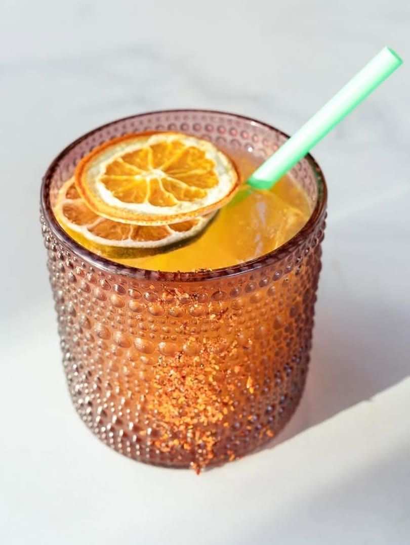 A vibrant orange cocktail in a textured glass, garnished with dried orange slices and a green straw.