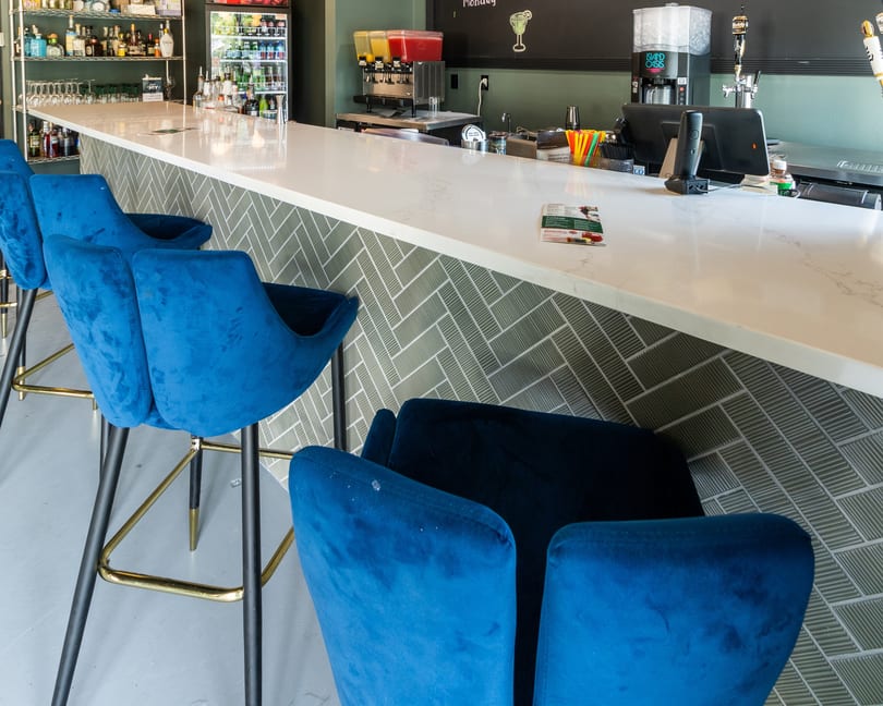 A modern bar with a white countertop, blue velvet bar stools, and a green tiled front.