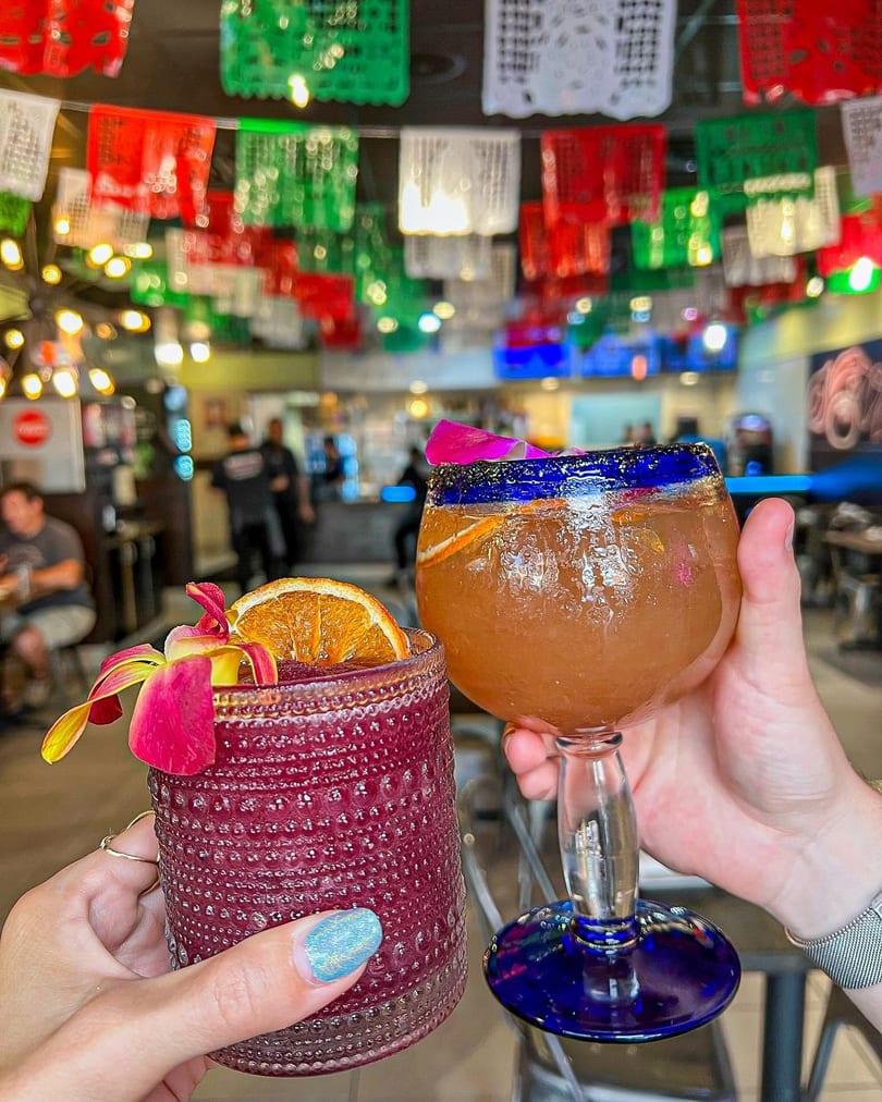 Two hands holding cocktails with orange slices and orchids, under colorful papel picado decorations.
